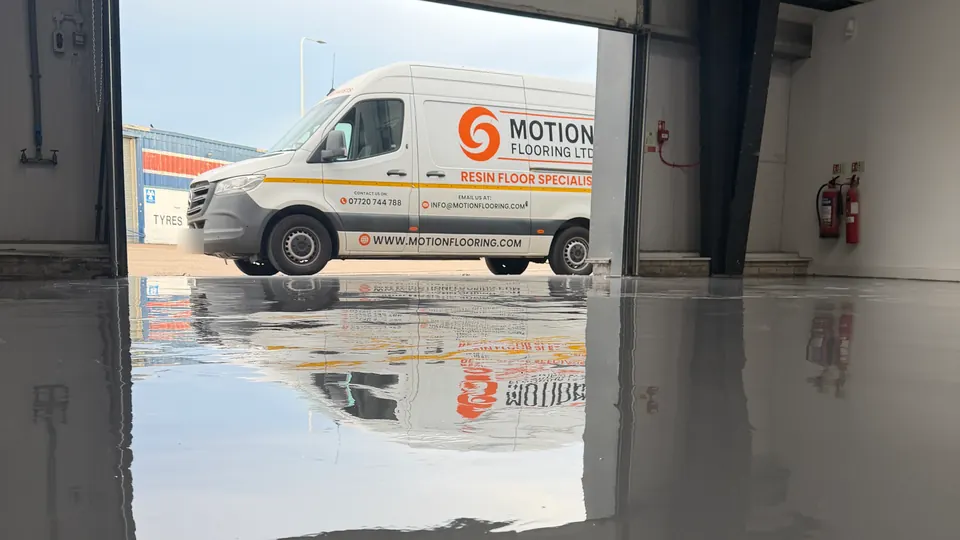 Freshly coated grey gloss floor in an industrial unit, with a Motion Flooring van reflected in the surface through the open roller door
